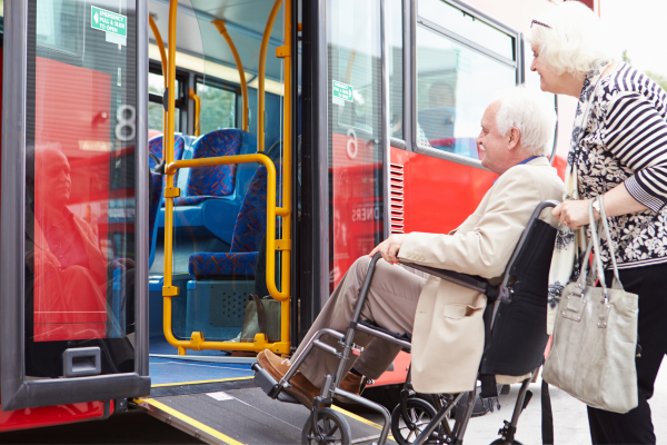 Image shows a bus with a ramp extended and a woman pushing a man on a wheelchair on to the bus